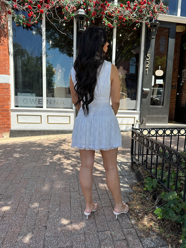 Woman wearing white lace mini dress with drop waist, v-neckline, and open back with tie detail. 