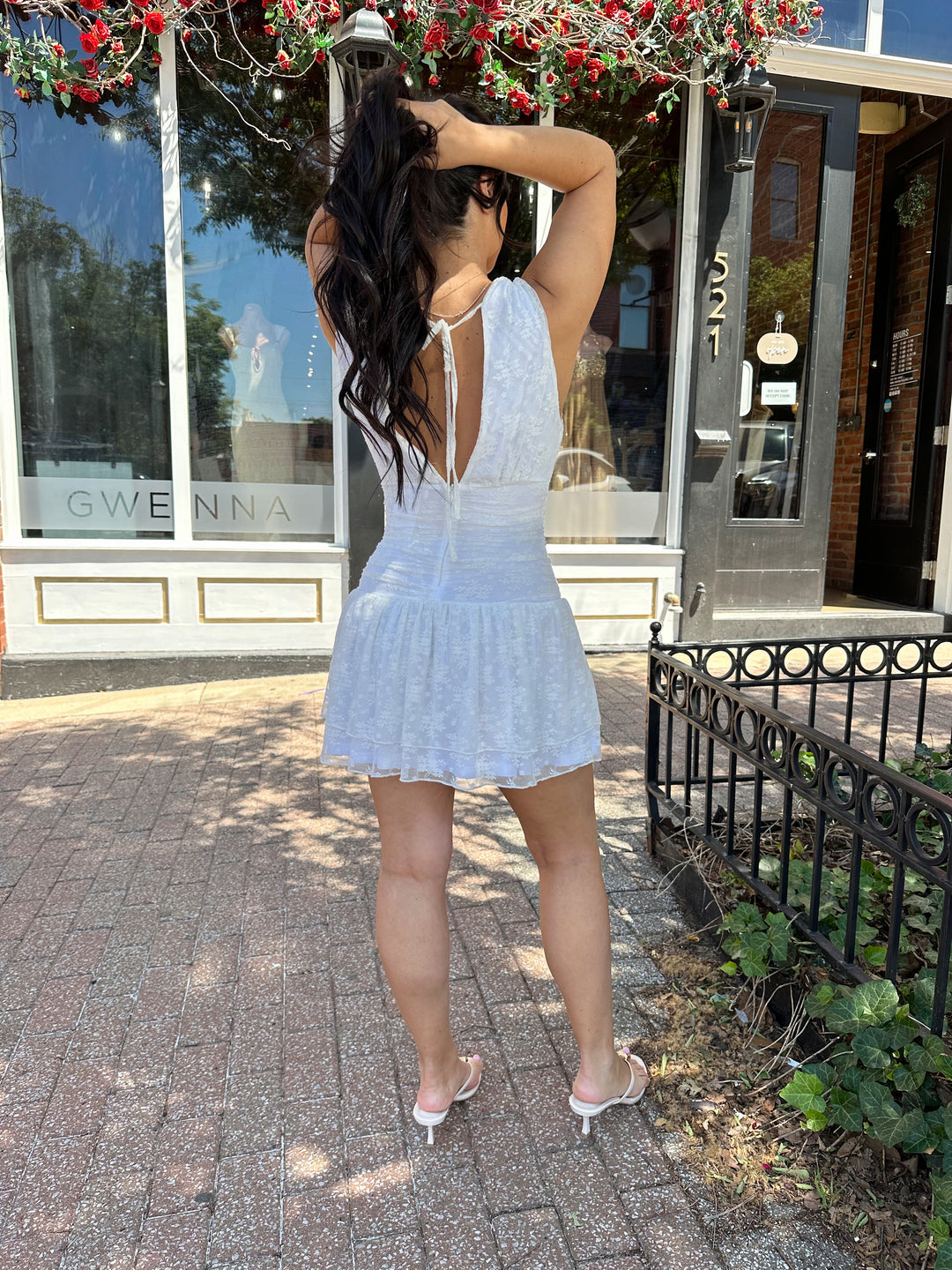 Woman wearing white lace mini dress with drop waist, v-neckline, and open back with tie detail. 