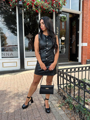 Woman wearing black leather mini dress with button detailing, belt loops, and functional pockets.