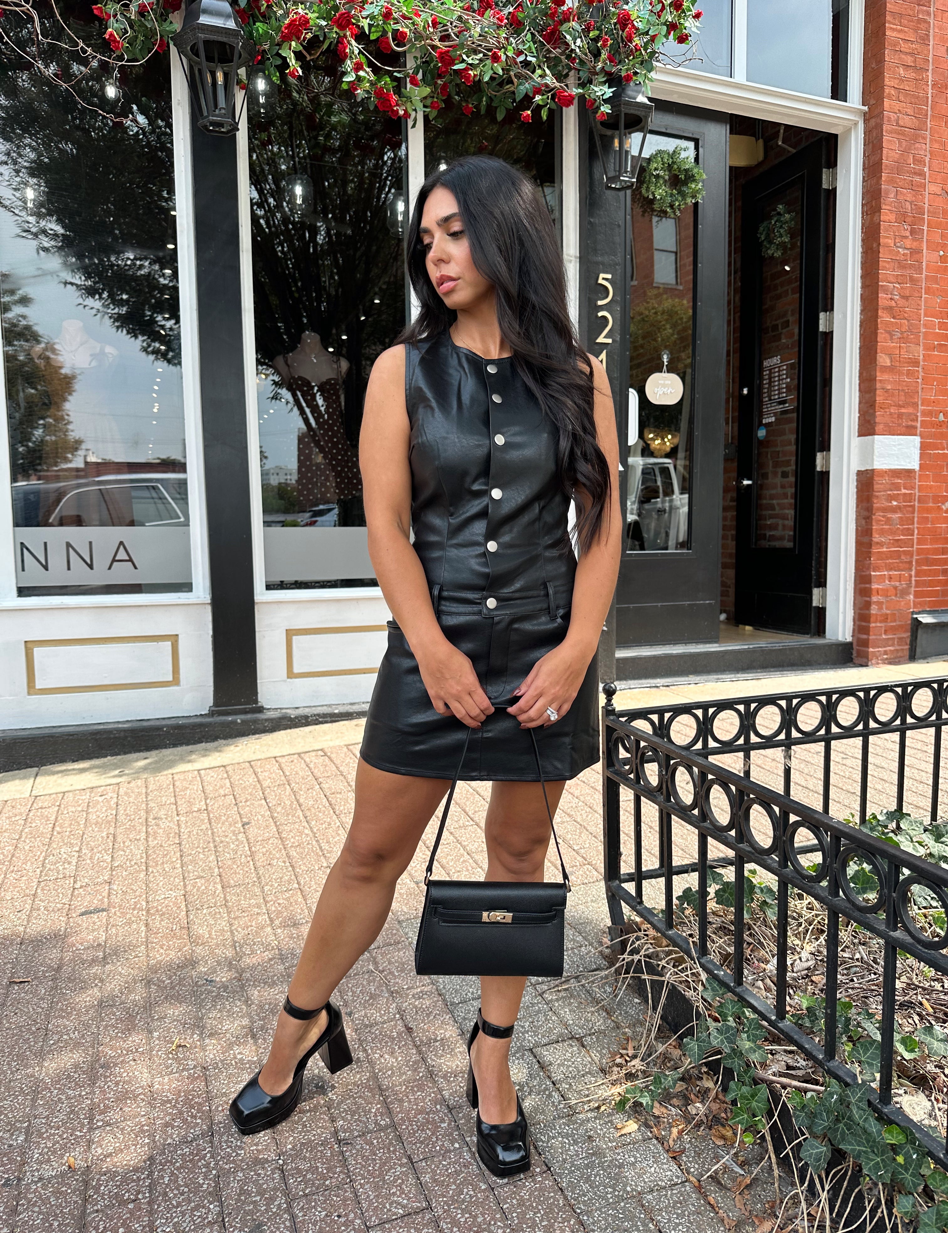 Woman wearing black leather mini dress with button detailing, belt loops, and functional pockets.