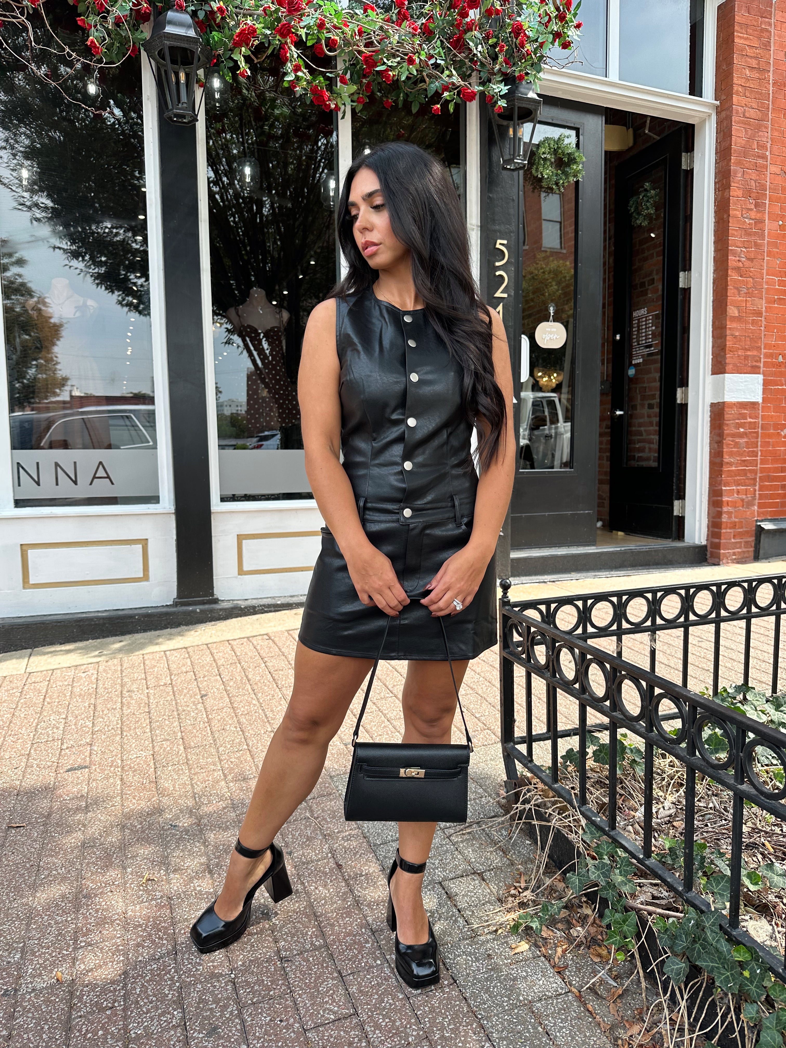 Woman wearing black leather mini dress with button detailing, belt loops, and functional pockets.