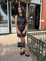 Woman wearing black leather mini dress with button detailing, belt loops, and functional pockets.
