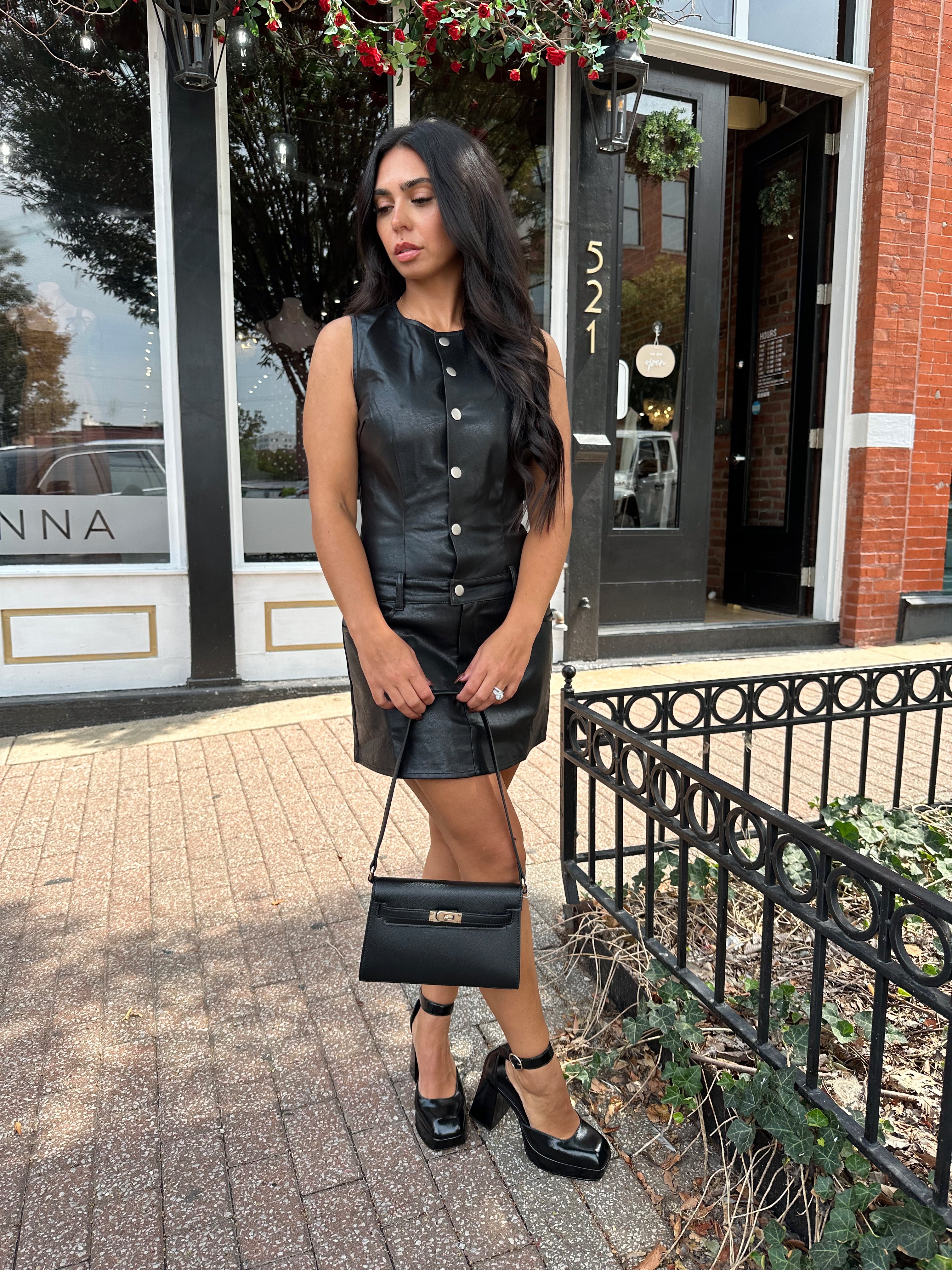 Woman wearing black leather mini dress with button detailing, belt loops, and functional pockets.