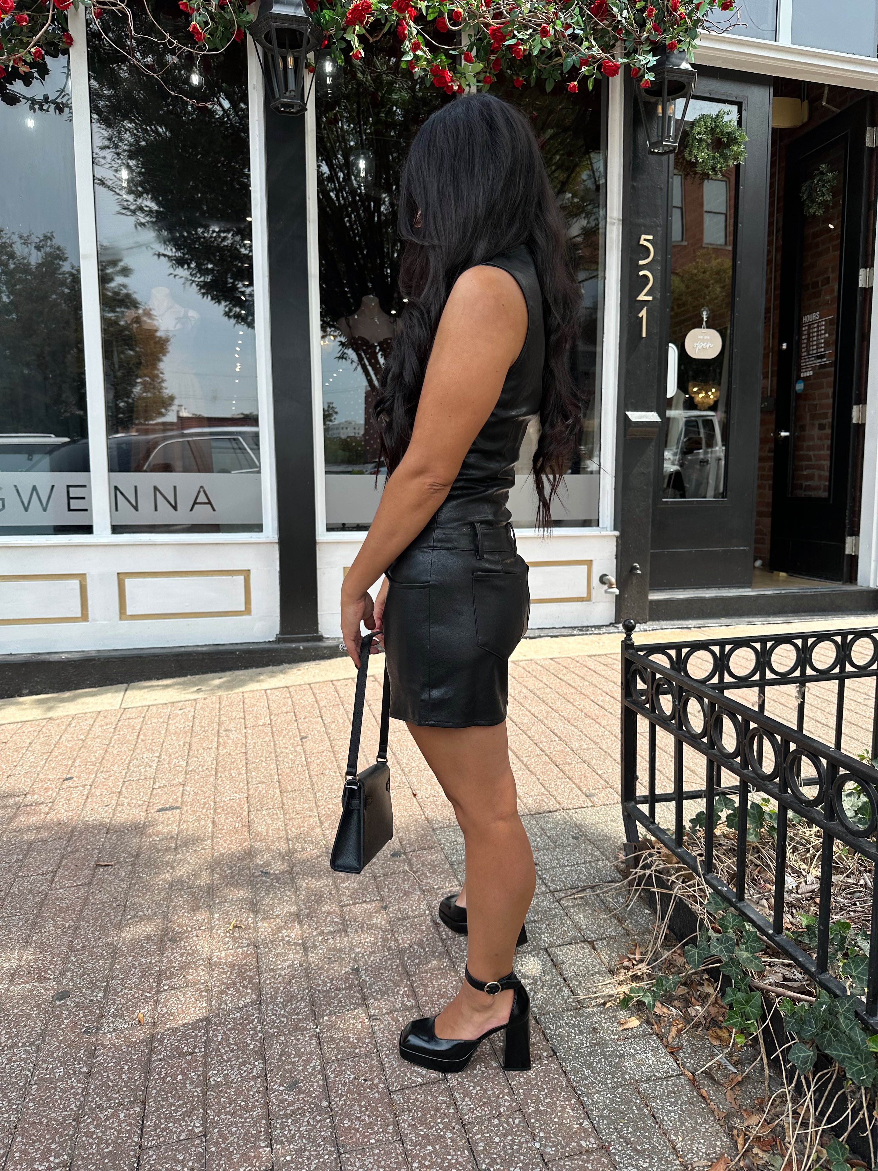 Woman wearing black leather mini dress with button detailing, belt loops, and functional pockets.