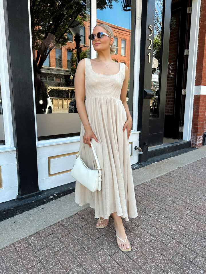 Woman wearing tan smocked midi dress with a fitted bodice and flowy skirt. Scoop neckline and functional pockets.