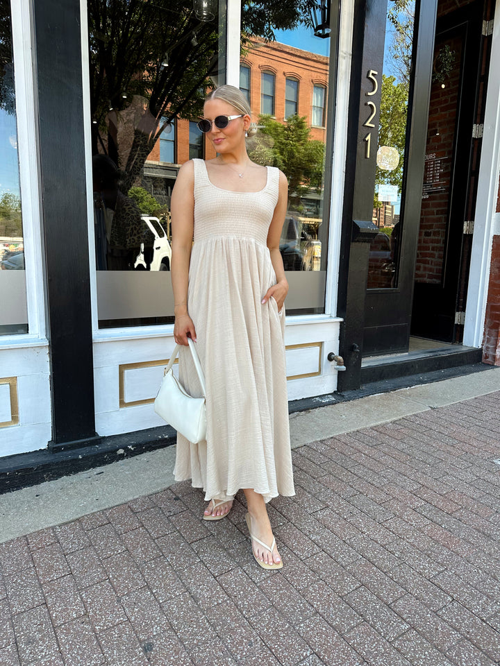 Woman wearing tan smocked midi dress with a fitted bodice and flowy skirt. Scoop neckline and functional pockets.