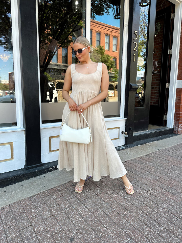 Woman wearing tan smocked midi dress with a fitted bodice and flowy skirt. Scoop neckline and functional pockets.