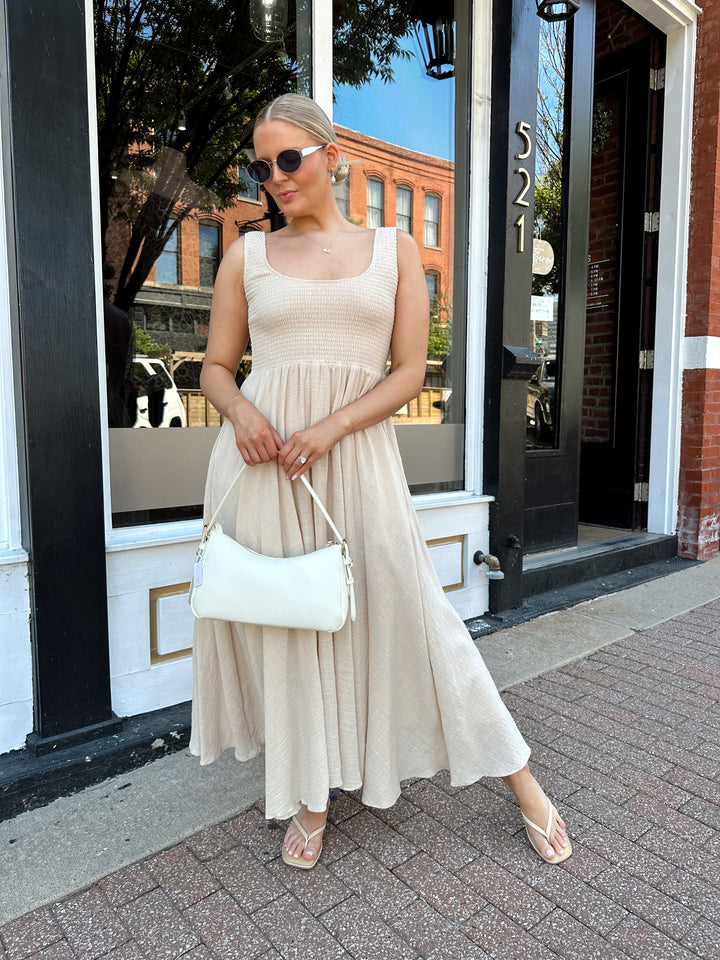 Woman wearing tan smocked midi dress with a fitted bodice and flowy skirt. Scoop neckline and functional pockets.