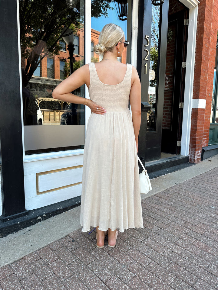 Woman wearing tan smocked midi dress with a fitted bodice and flowy skirt. Scoop neckline and functional pockets.