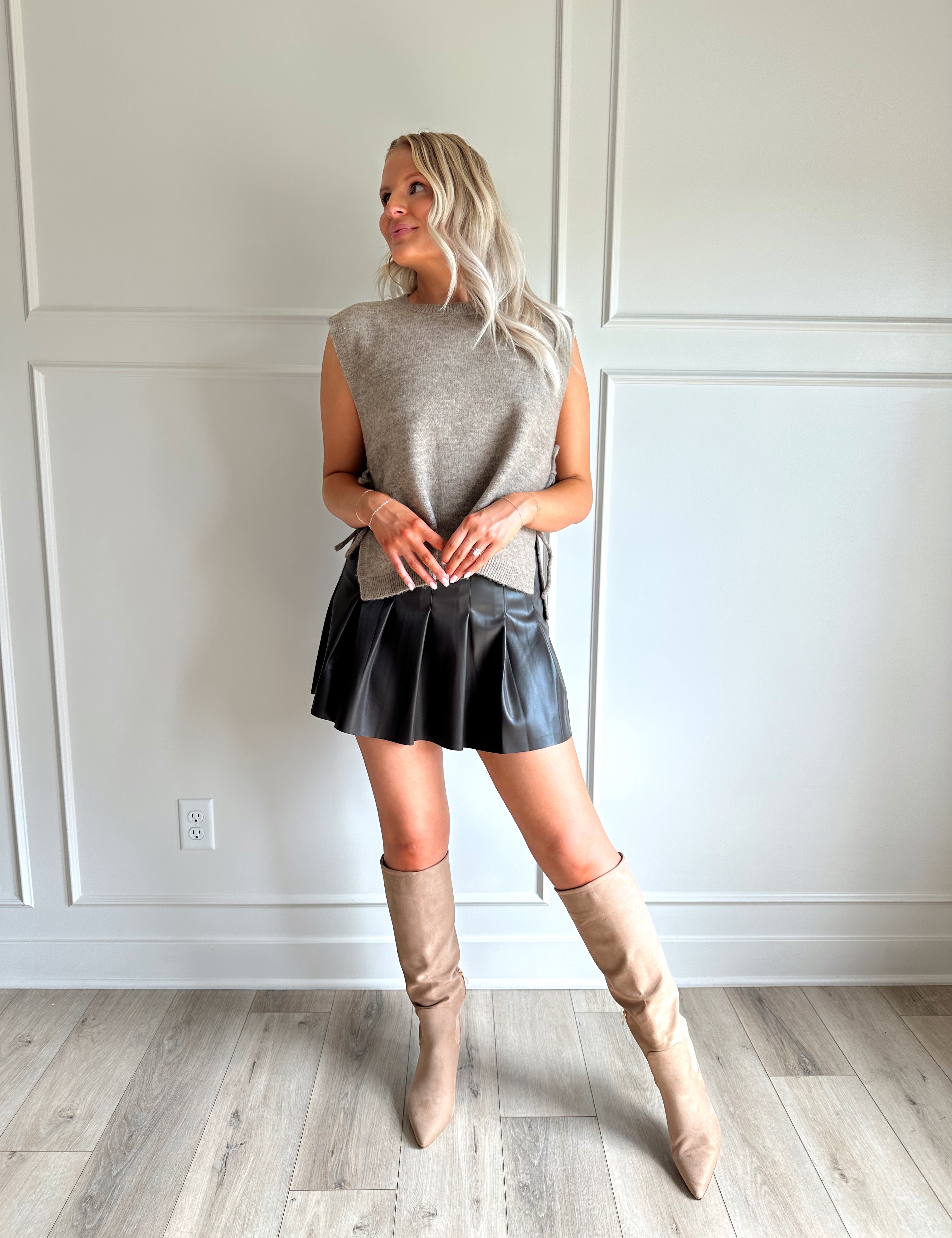 Woman wearing a brown sleeveless sweater top with tie sides and a brown pleated mini skirt.