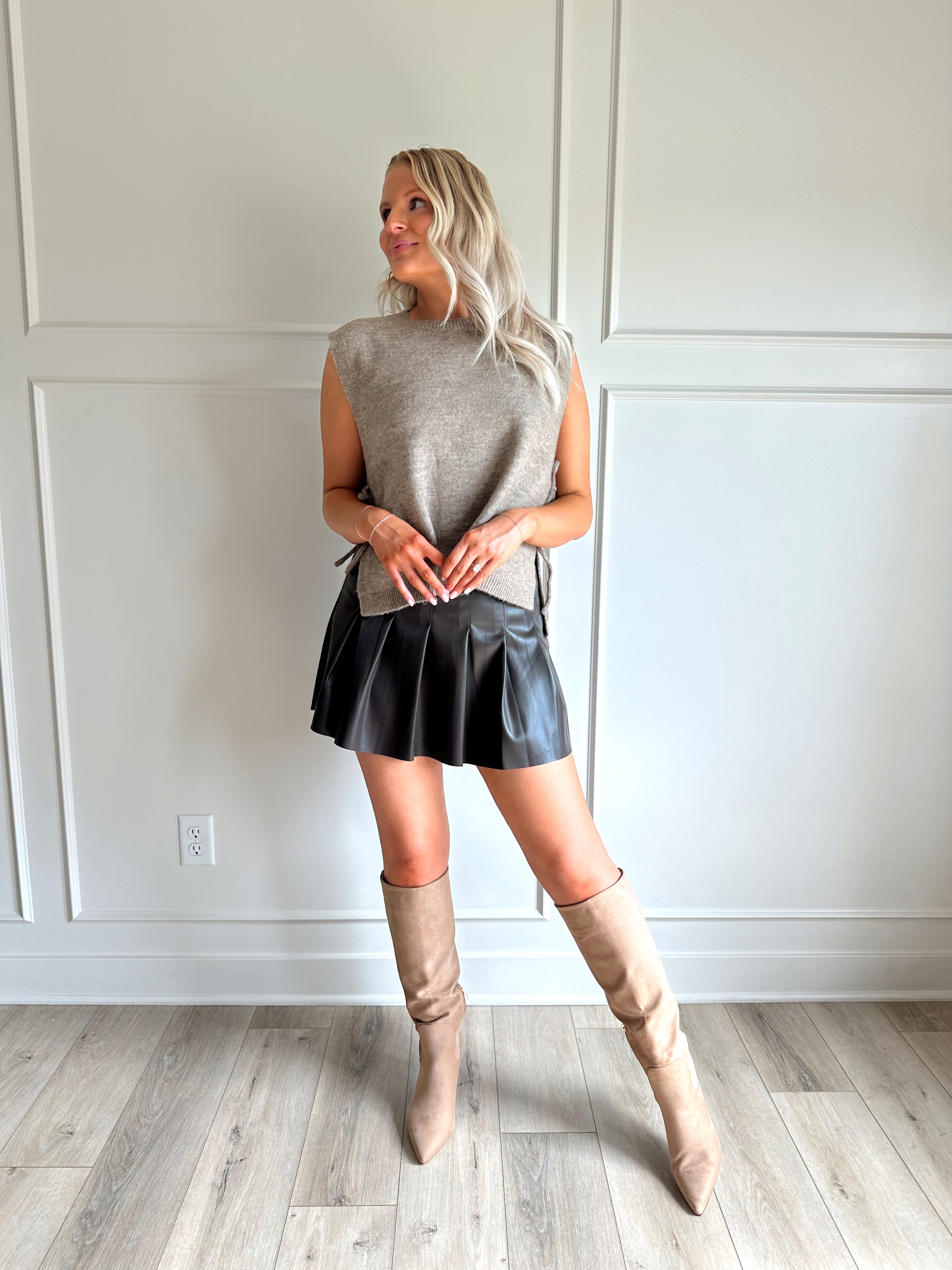 Woman wearing a brown sleeveless sweater top with tie sides and a brown pleated mini skirt.