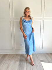 Woman wearing a light blue satin midi dress with ruffle trim along neckline. 