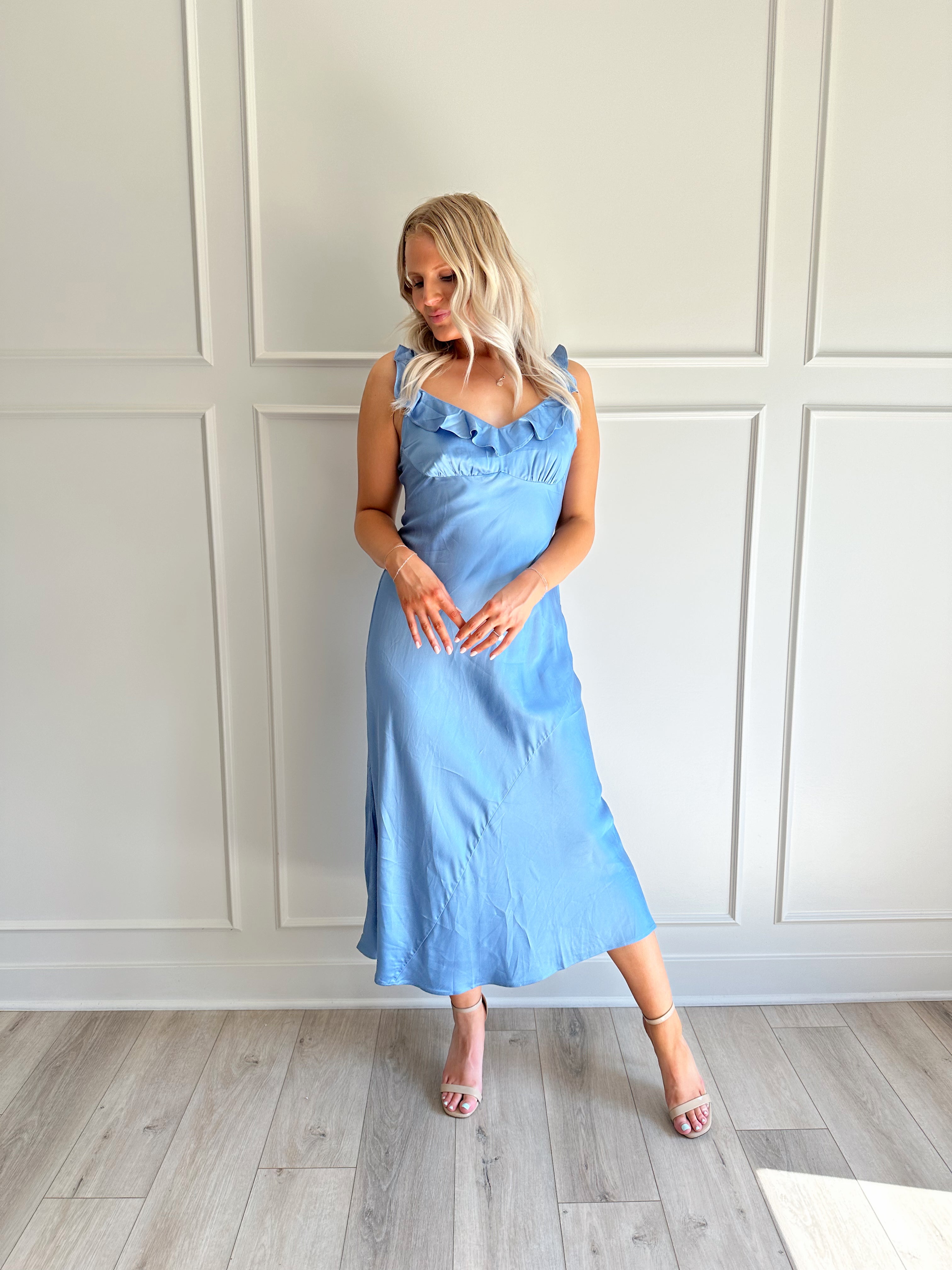Woman wearing a light blue satin midi dress with ruffle trim along neckline. 