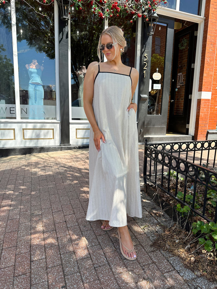 Woman wearing neutral pinstripe maxi dress, with adjustable spaghetti straps. 