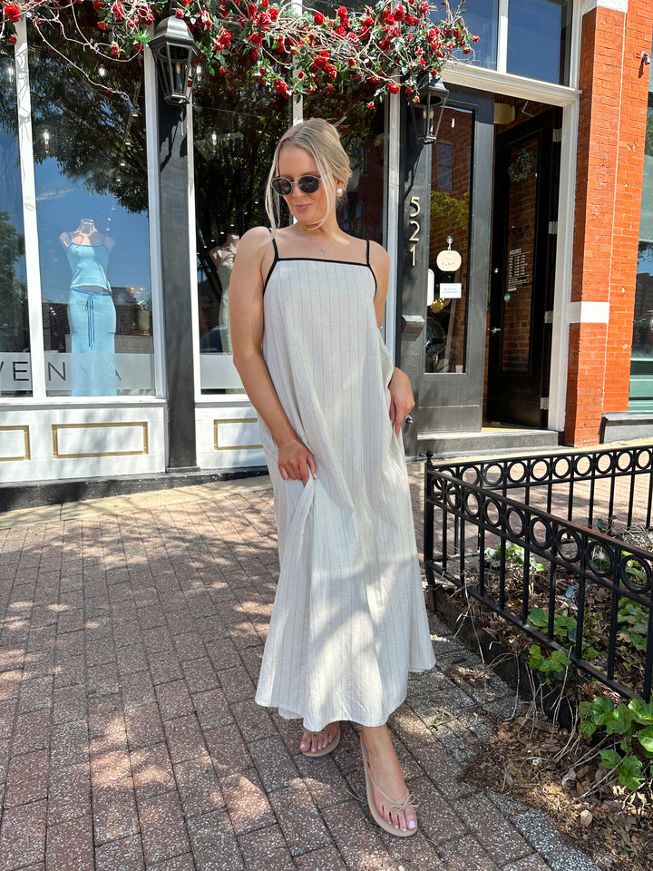 Woman wearing neutral pinstripe maxi dress, with adjustable spaghetti straps. 