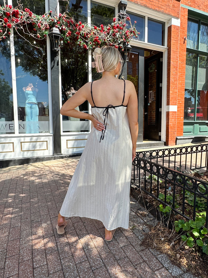 Woman wearing neutral pinstripe maxi dress, with adjustable spaghetti straps. 