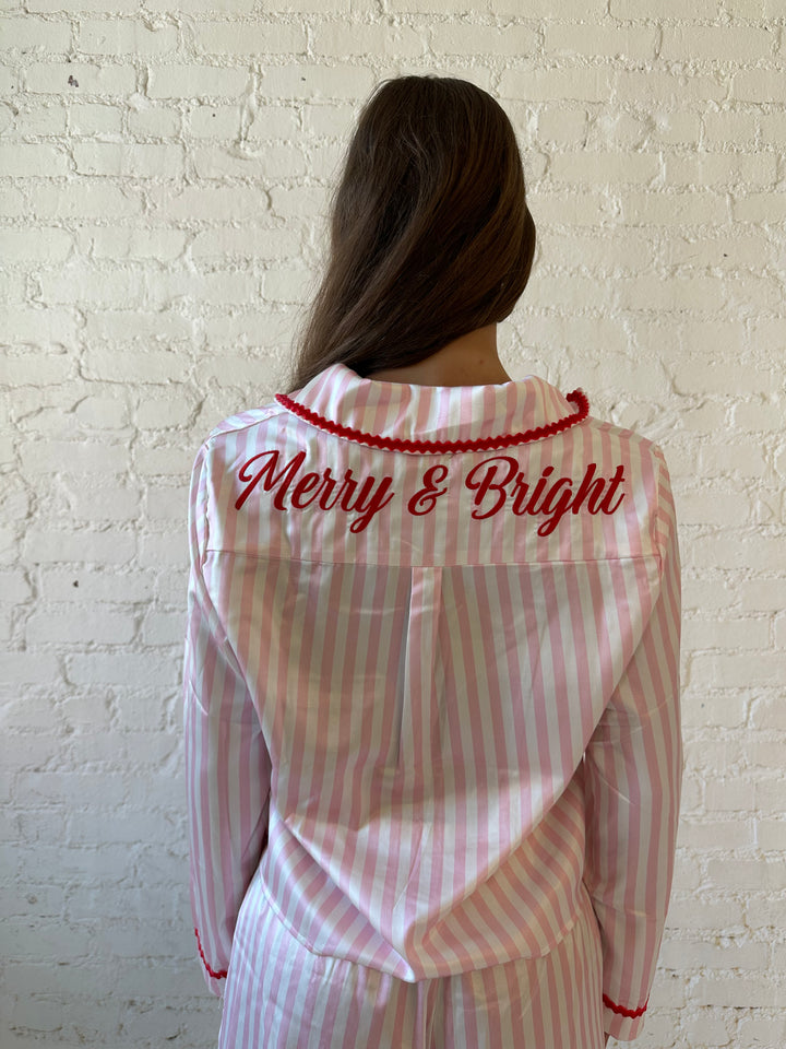 Woman wearing light pink and white striped pajamas with red trim and red Merry and Bright wording on the back. 