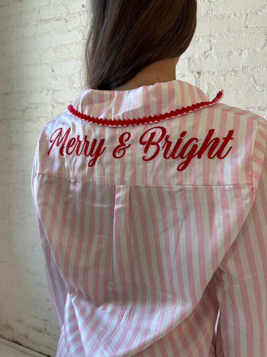 Woman wearing light pink and white striped pajamas with red trim and red Merry and Bright wording on the back. 