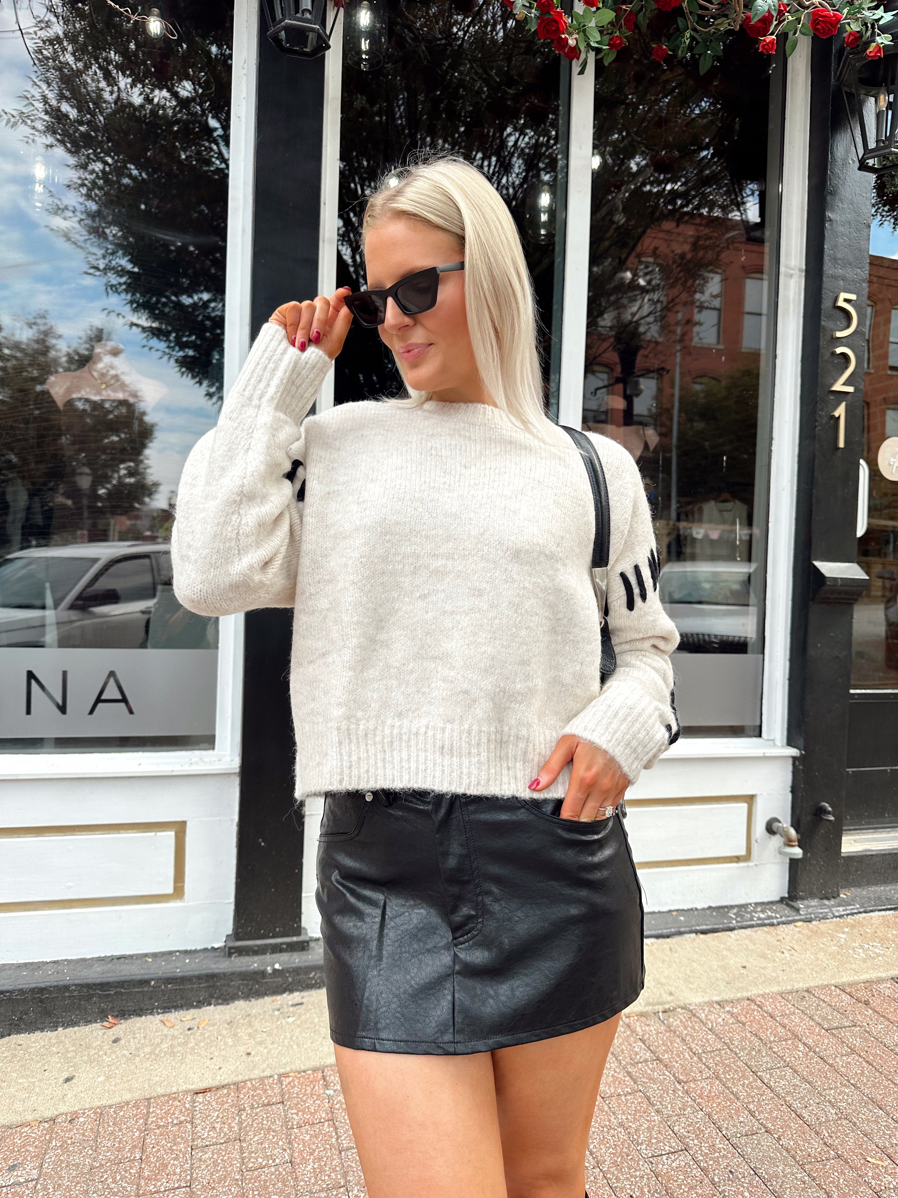 Woman wearing oatmeal knit sweater with black stitch sleeve detailing. 