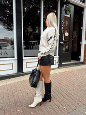 Woman wearing oatmeal knit sweater with black stitch sleeve detailing. 