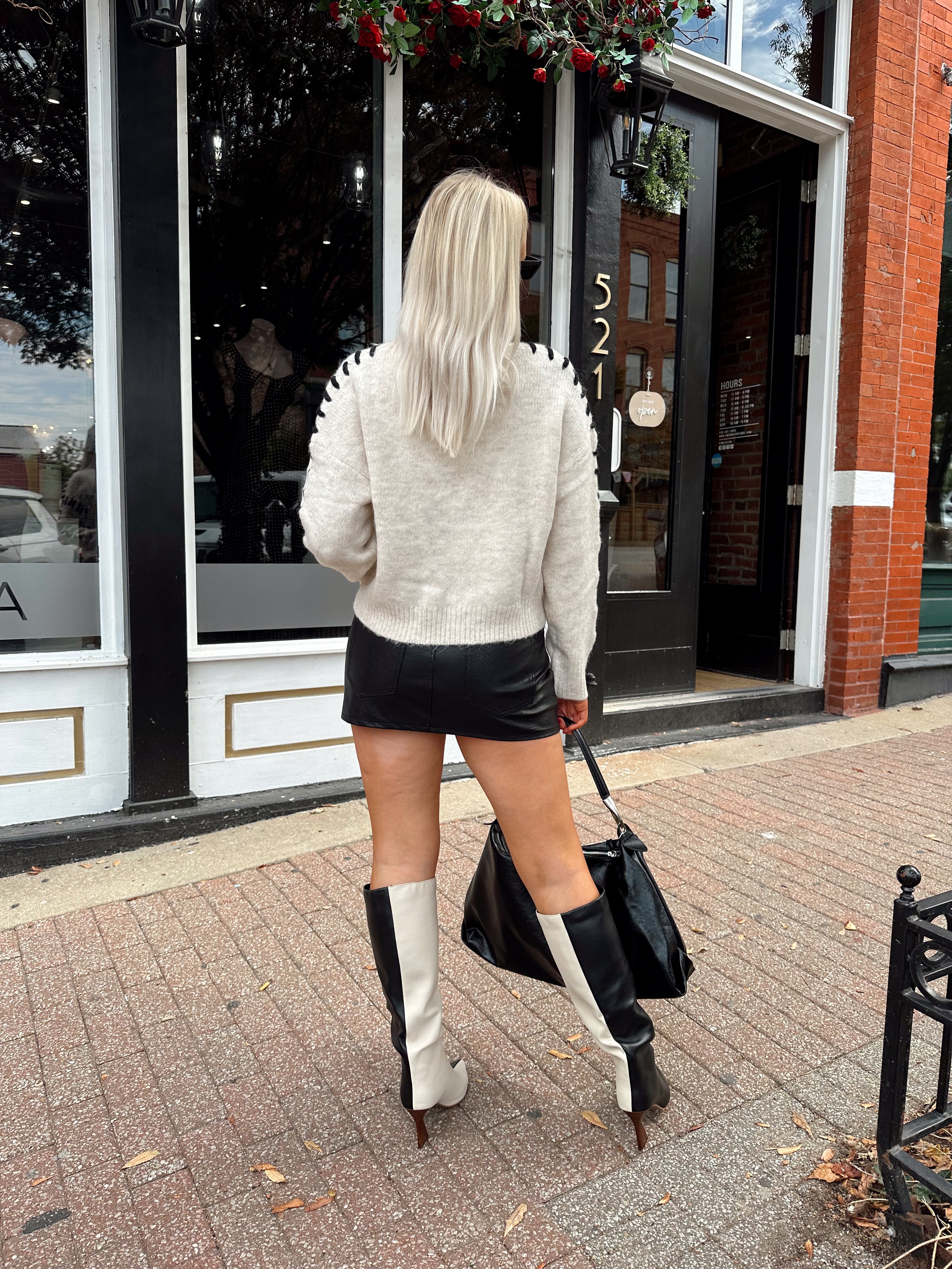 Woman wearing oatmeal knit sweater with black stitch sleeve detailing. 