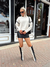 Woman wearing oatmeal knit sweater with black stitch sleeve detailing. 