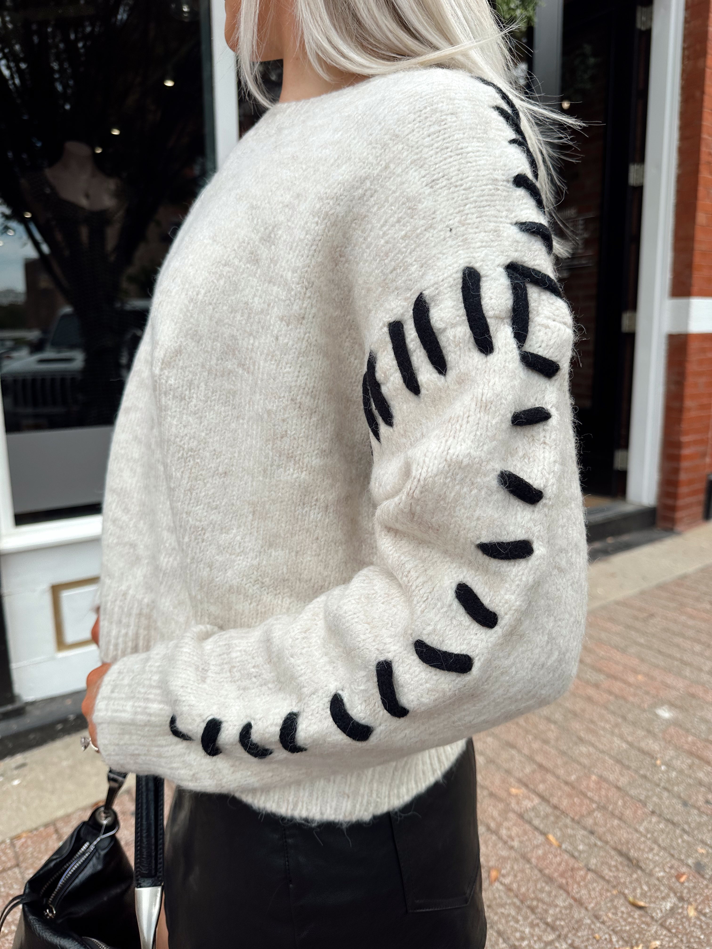 Woman wearing oatmeal knit sweater with black stitch sleeve detailing. 
