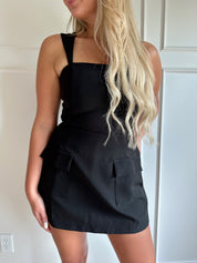 Woman wearing a black tailored mini dress with pocket flaps and built in shorts. 