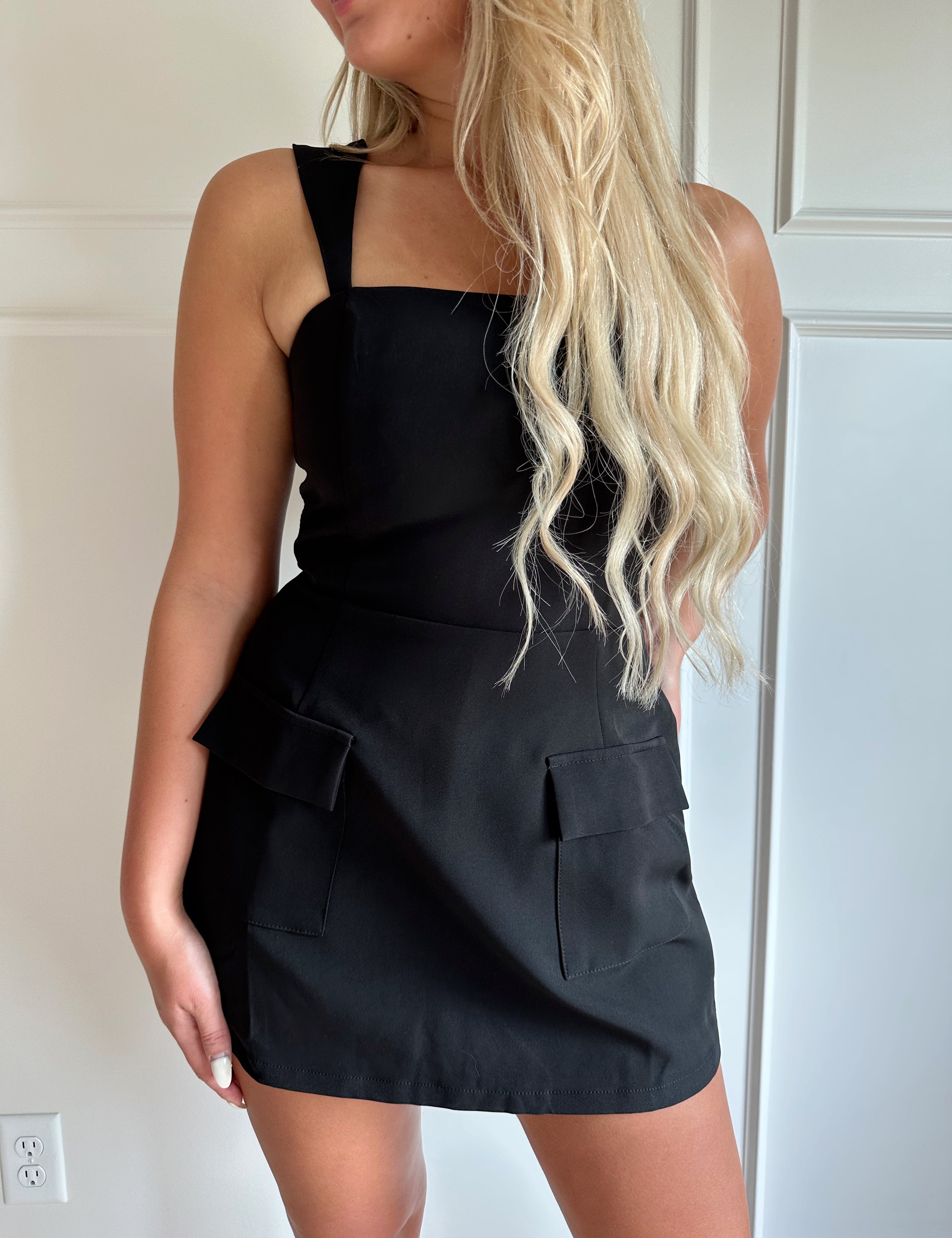 Woman wearing a black tailored mini dress with pocket flaps and built in shorts. 