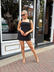 Woman wearing black tailored mini dress with adjustable straps and built in shorts.