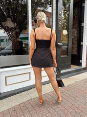 Woman wearing black tailored mini dress with adjustable straps and built in shorts.