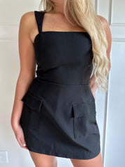 Woman wearing a black tailored mini dress with pocket flaps and built in shorts. 