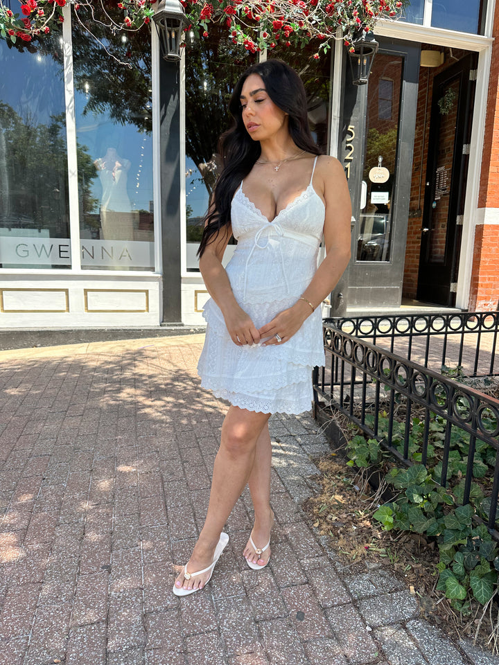 Woman wearing white lace tiered mini dress with adjustable spaghetti straps and tie detail. 
