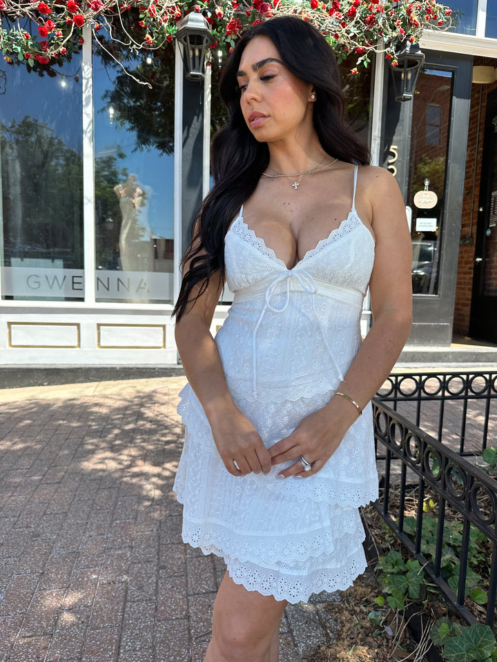 Woman wearing white lace tiered mini dress with adjustable spaghetti straps and tie detail. 