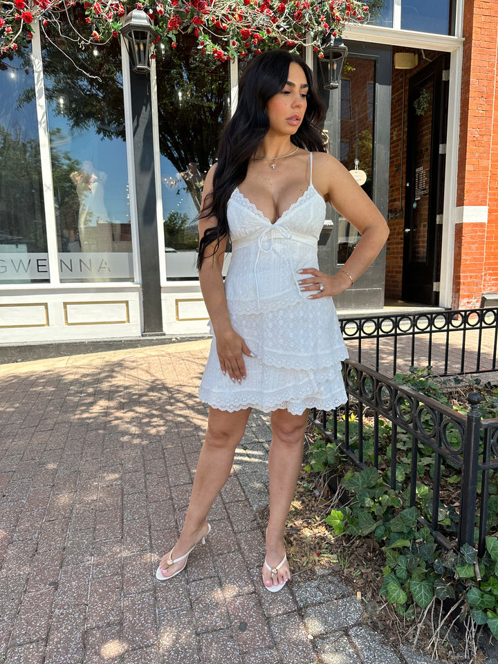 Woman wearing white lace tiered mini dress with adjustable spaghetti straps and tie detail. 