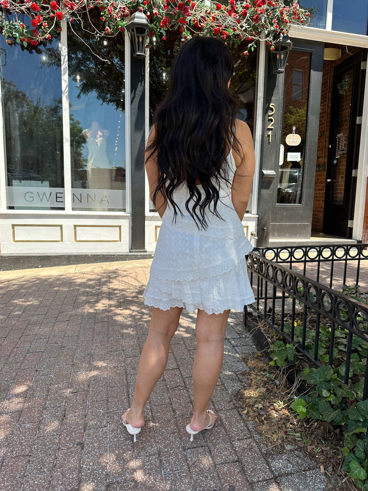 Woman wearing white lace tiered mini dress with adjustable spaghetti straps and tie detail. 