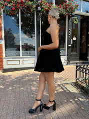 Woman in a black dress standing on a sidewalk with decorative plants above.