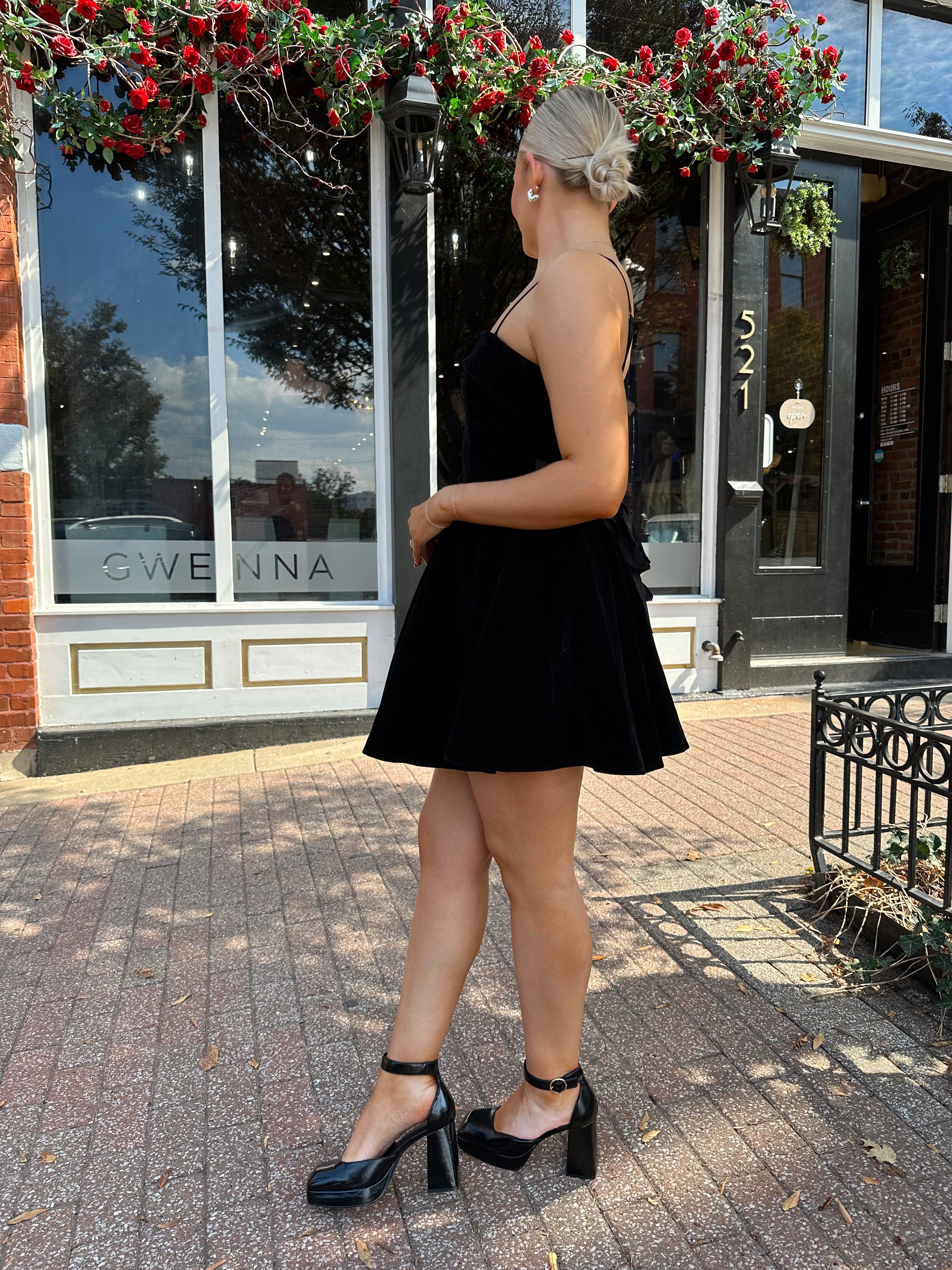 Woman in a black dress standing on a sidewalk with decorative plants above.