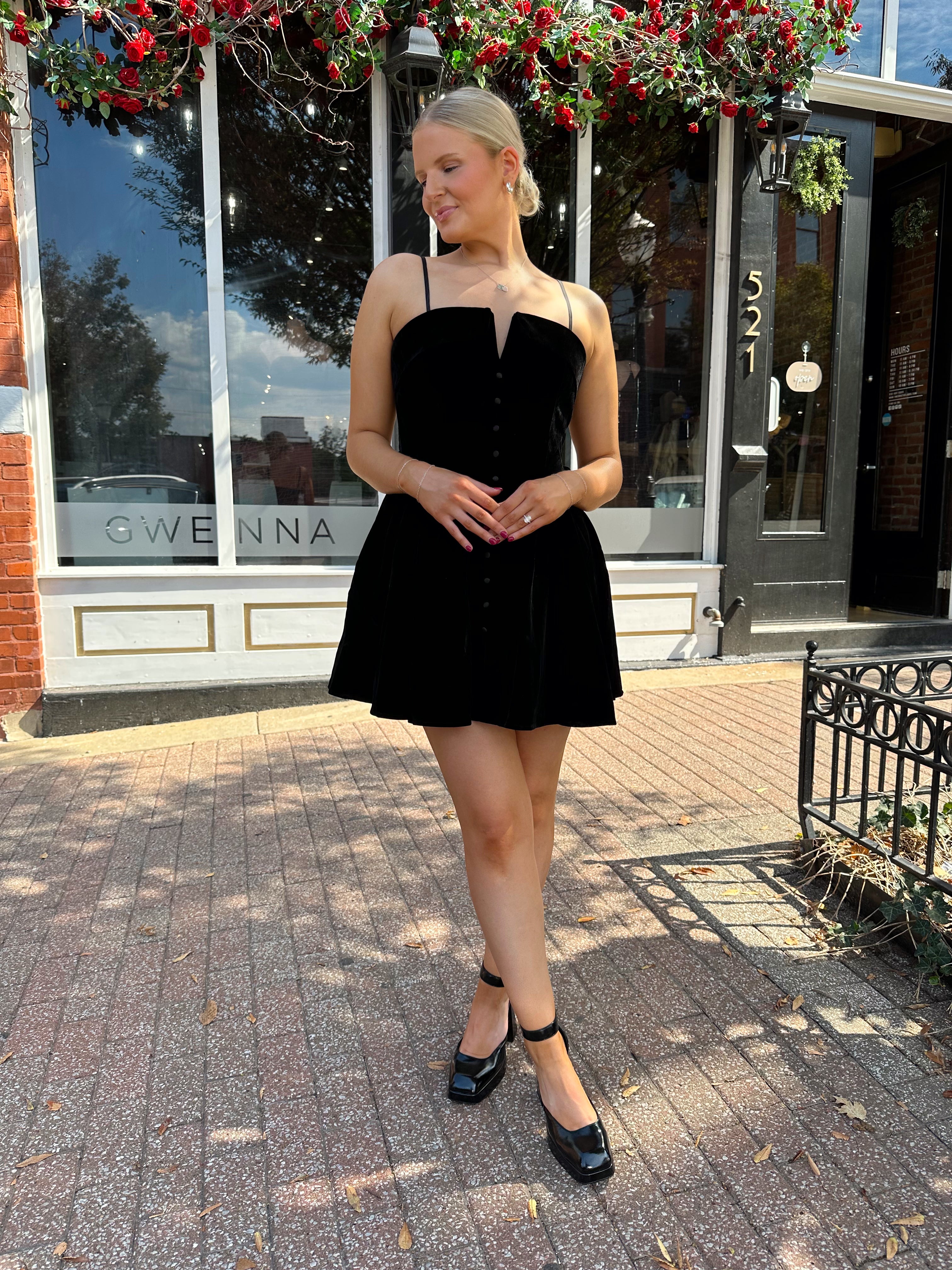 Woman in a black dress standing on a sidewalk in front of a building with a sign that reads 'Gwen'.