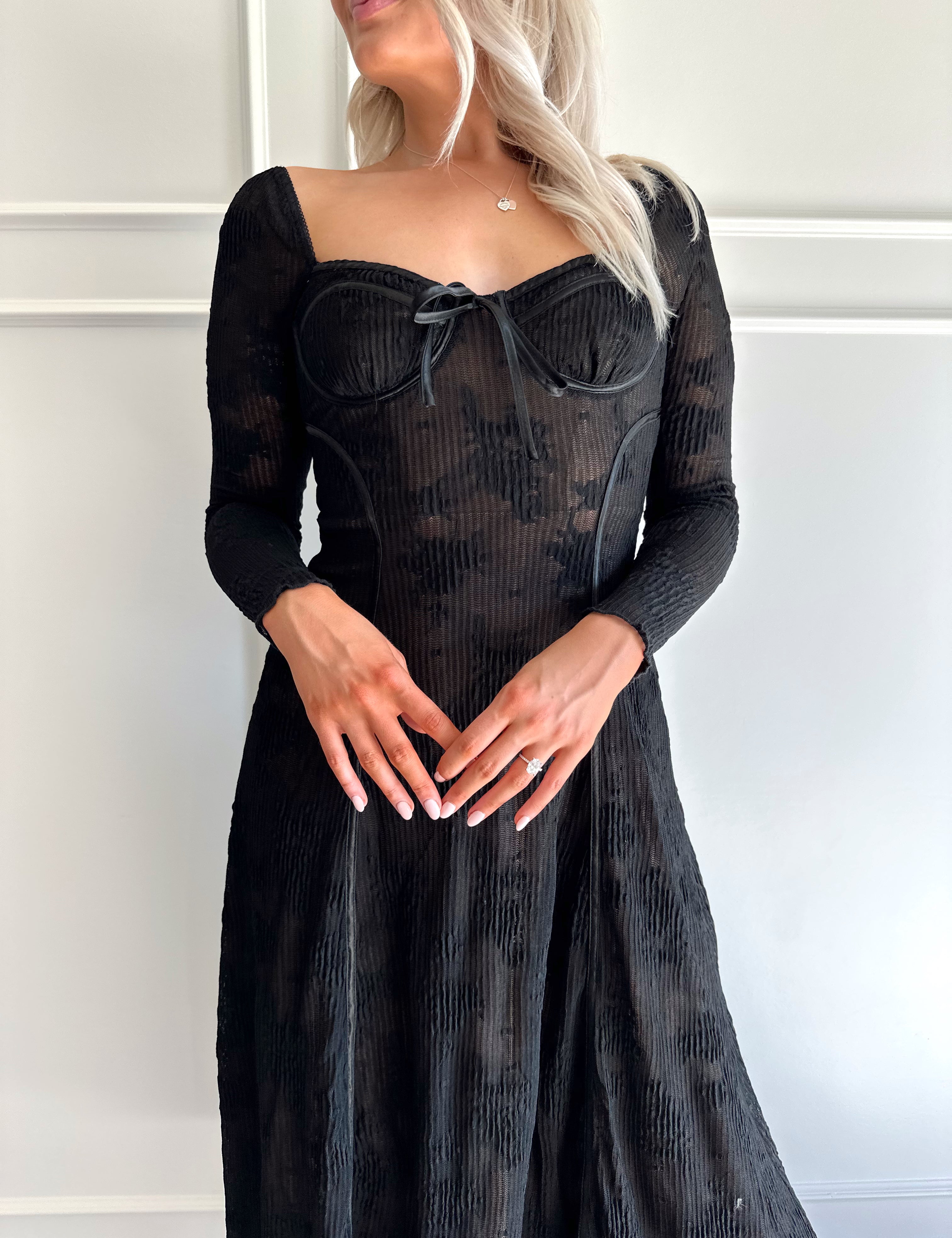 Woman wearing a black lace midi dress with long sleeves, tie detail, and flowy bottom.