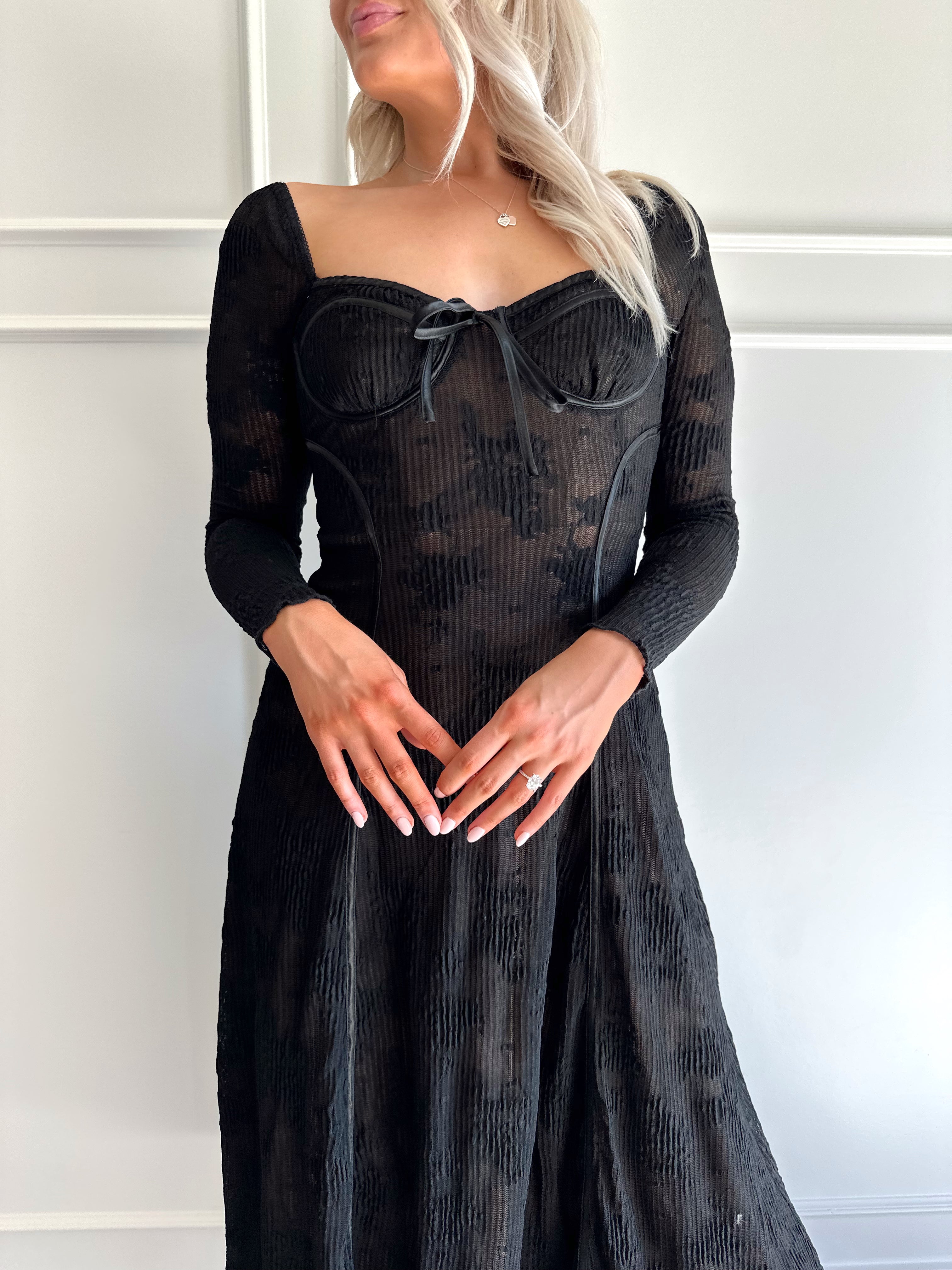 Woman wearing a black lace midi dress with long sleeves, tie detail, and flowy bottom.