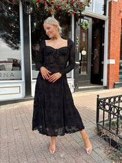 Woman wearing long sleeve black lace midi dress with tan underlay. 