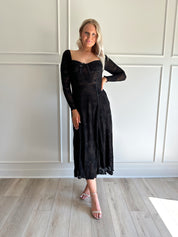 Woman wearing a black lace midi dress with long sleeves, tie detail, and flowy bottom.