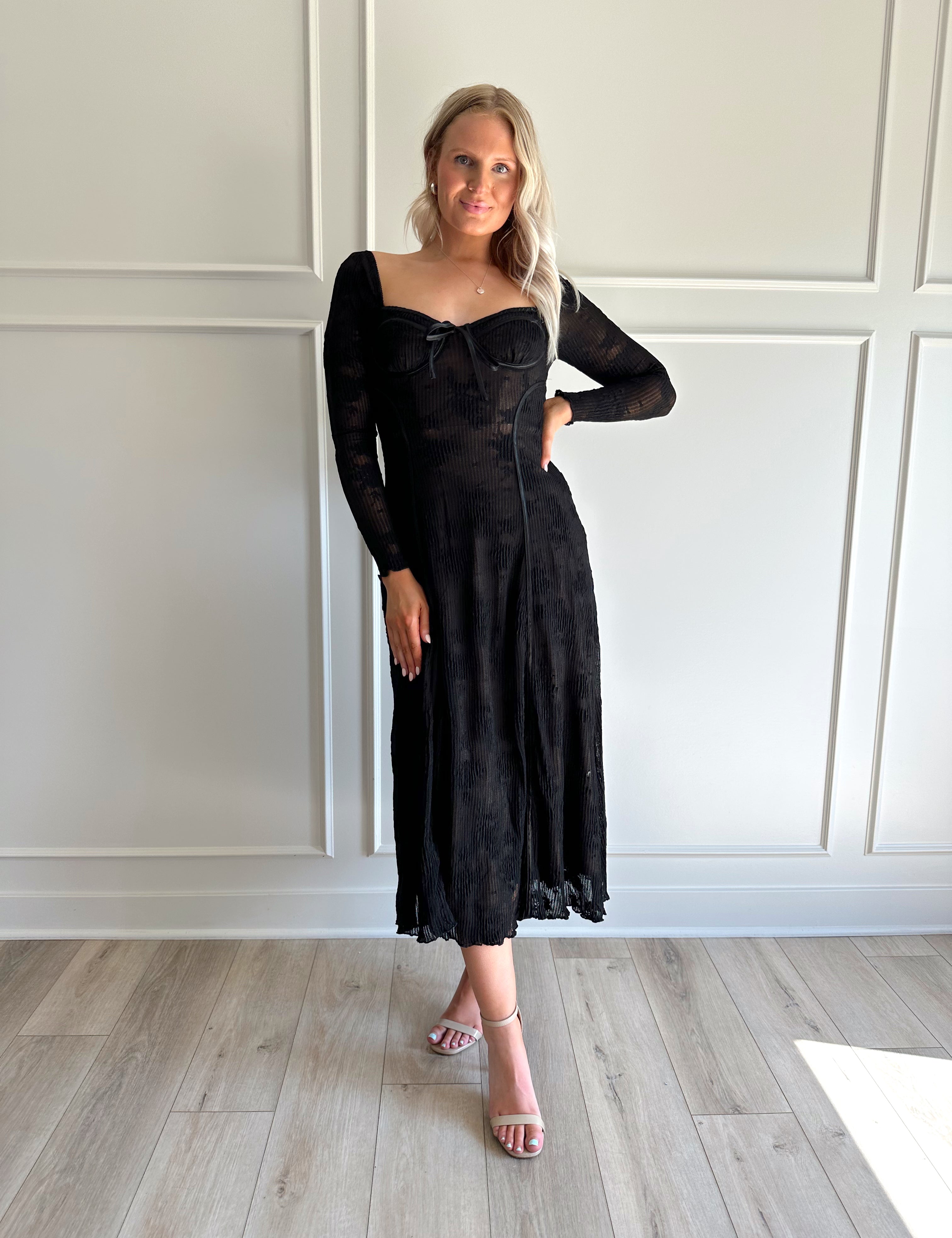 Woman wearing a black lace midi dress with long sleeves, tie detail, and flowy bottom.