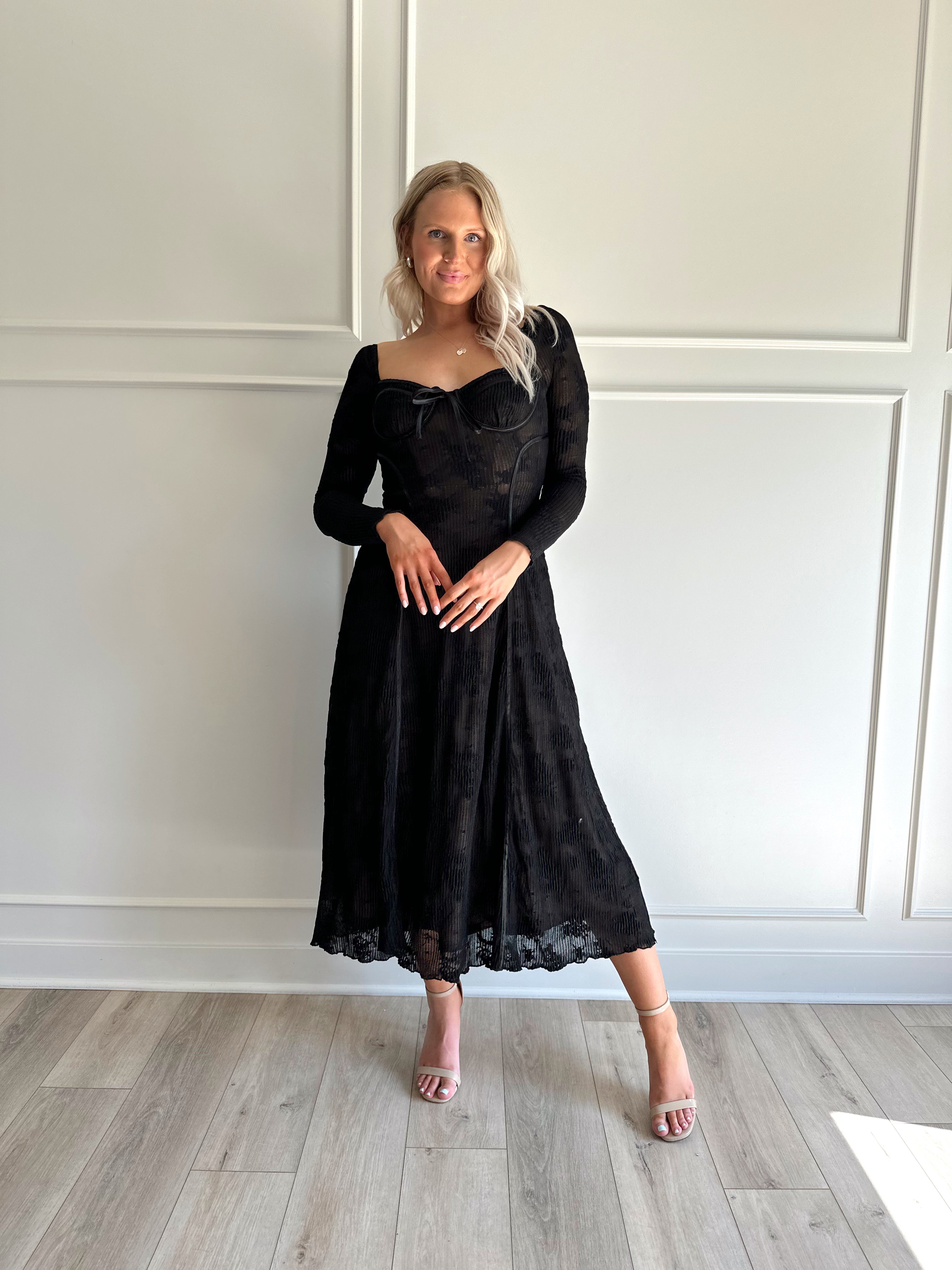 Woman wearing a black lace midi dress with long sleeves, tie detail, and flowy bottom.