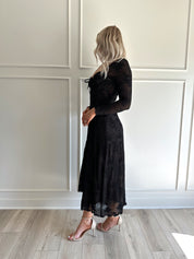 Woman wearing a black lace midi dress with long sleeves, tie detail, and flowy bottom.