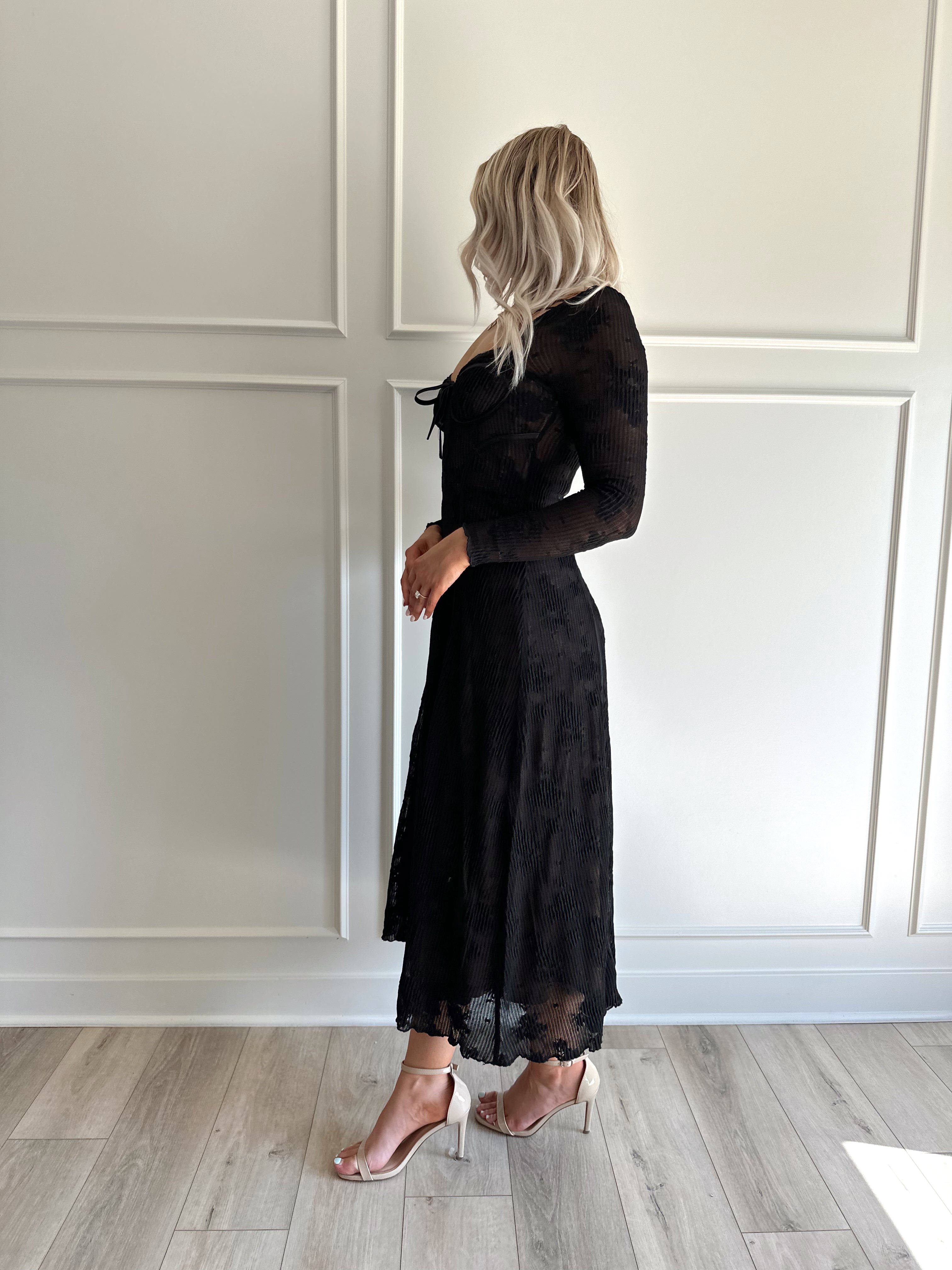 Woman wearing a black lace midi dress with long sleeves, tie detail, and flowy bottom.