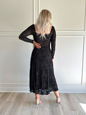 Woman wearing a black lace midi dress with long sleeves, tie detail, and flowy bottom.