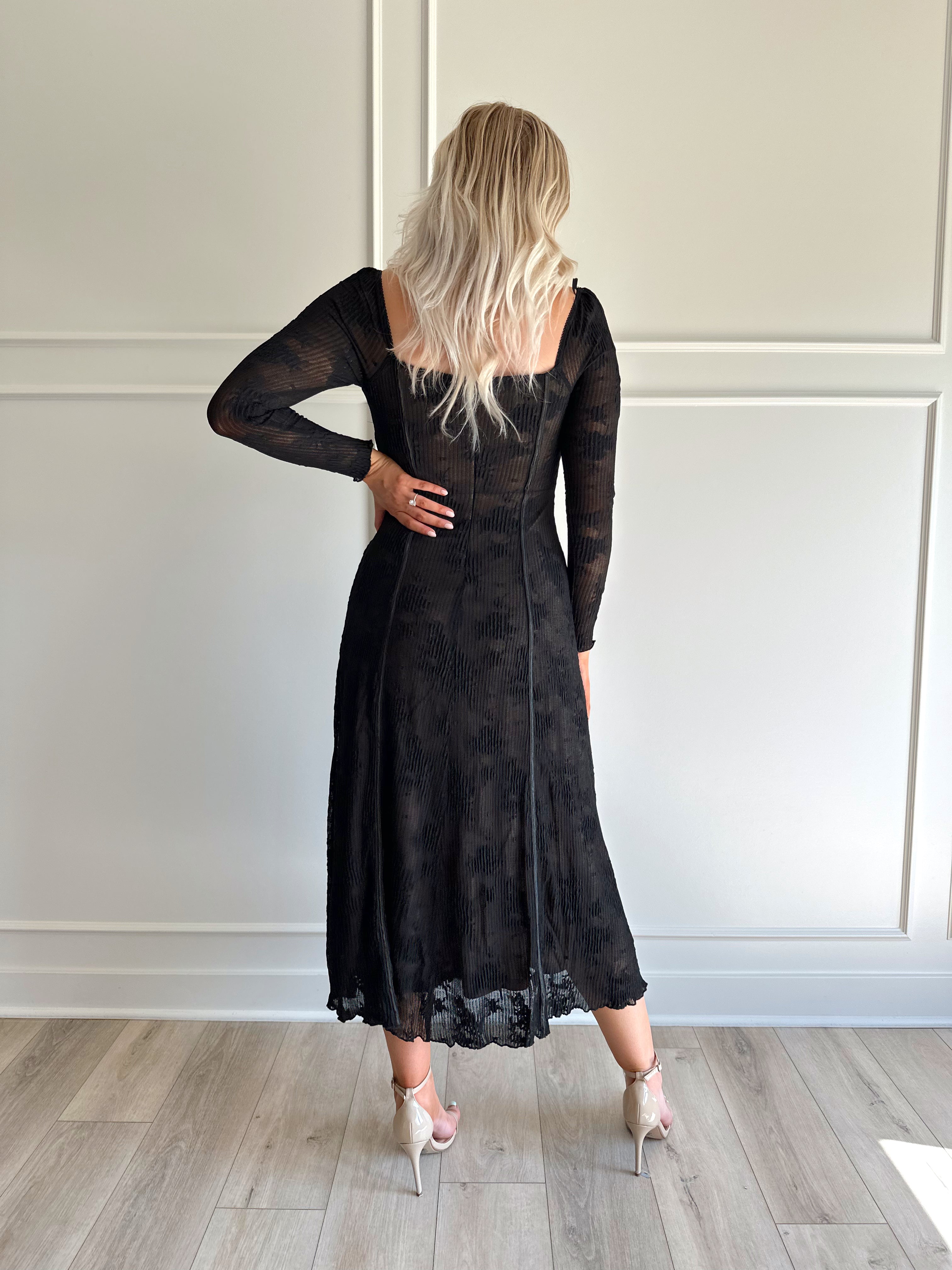 Woman wearing a black lace midi dress with long sleeves, tie detail, and flowy bottom.