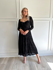 Woman wearing a black lace midi dress with long sleeves, tie detail, and flowy bottom.